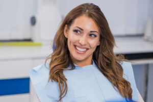 Smiling woman with long brown hair sitting in a dental clinic, wearing a light blue dental bib, looking off to the side.
